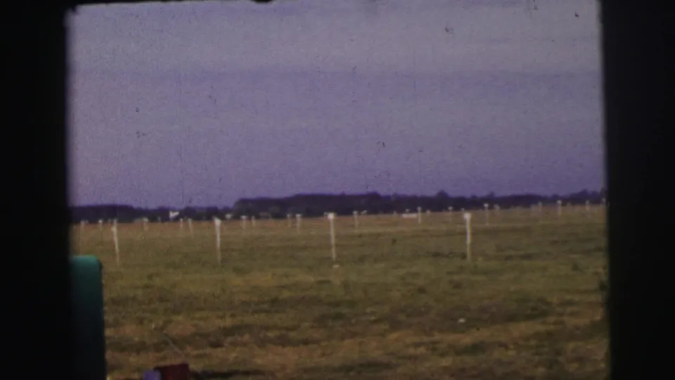 Vehicle-framed 1954 airfield view in Le Mans, showing grass, field markers, a distant tree line, and a green foreground object.