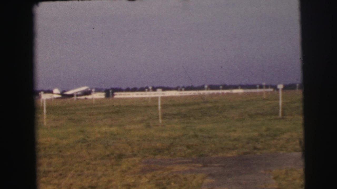 A white passenger plane appears near low airport structures on the Le Mans grass airfield, framed by dark 8mm borders.