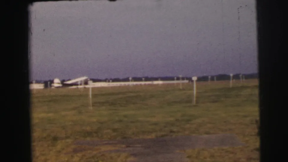 The Le Mans airfield scene shows a white passenger aircraft near low buildings, with grass foreground and white posts marking the field.