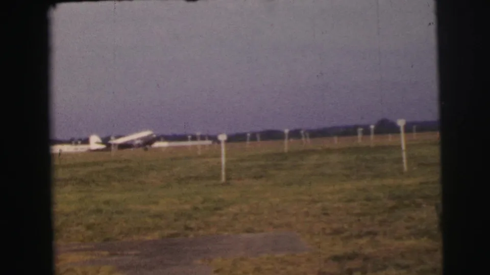A white passenger plane is seen on the Le Mans grass airfield under a muted sky, with white field posts and distant trees behind it.