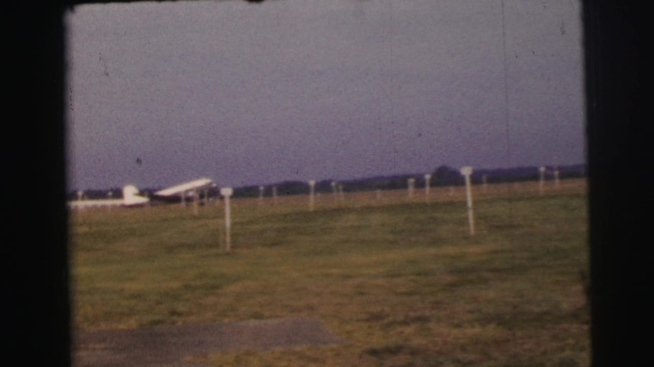 Small white propeller plane parked on a grassy airfield with marker poles and an overcast sky.