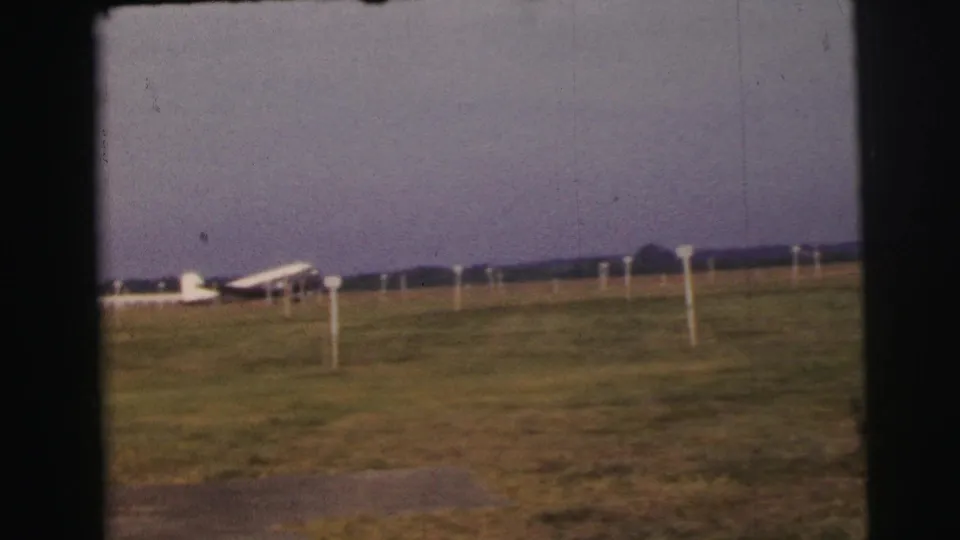 1954 Le Mans home-movie frame showing a small white propeller plane on grass beyond white marker poles under a muted sky.