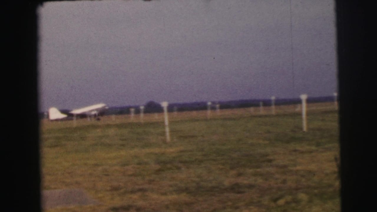 1954 Le Mans 8mm frame with a distant white propeller plane on a grassy field and simple airfield markers.