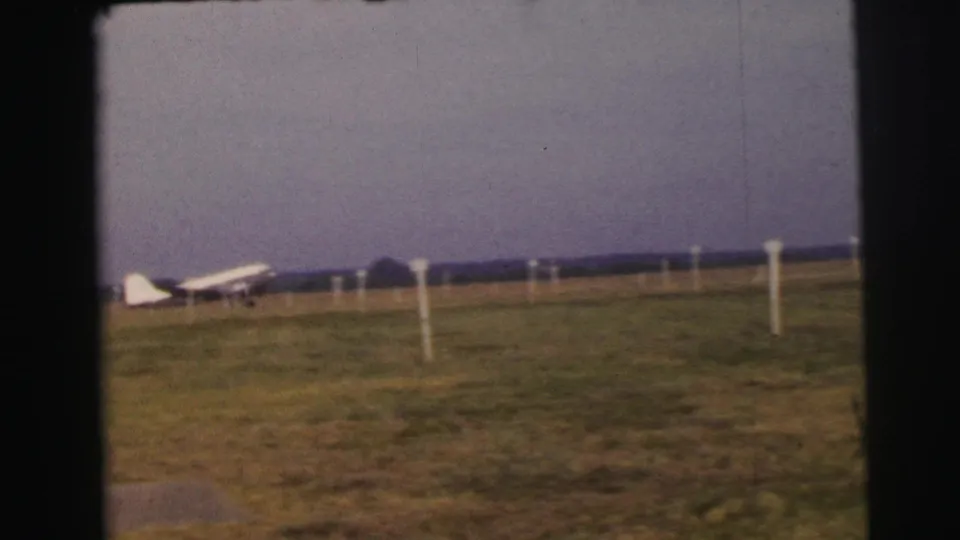 1954 Le Mans 8mm frame with a distant white propeller plane on a grassy field and simple airfield markers.