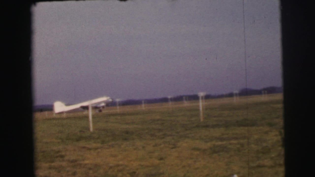 1954 Le Mans home-movie frame showing a small white airplane near a grass runway with white boundary posts.