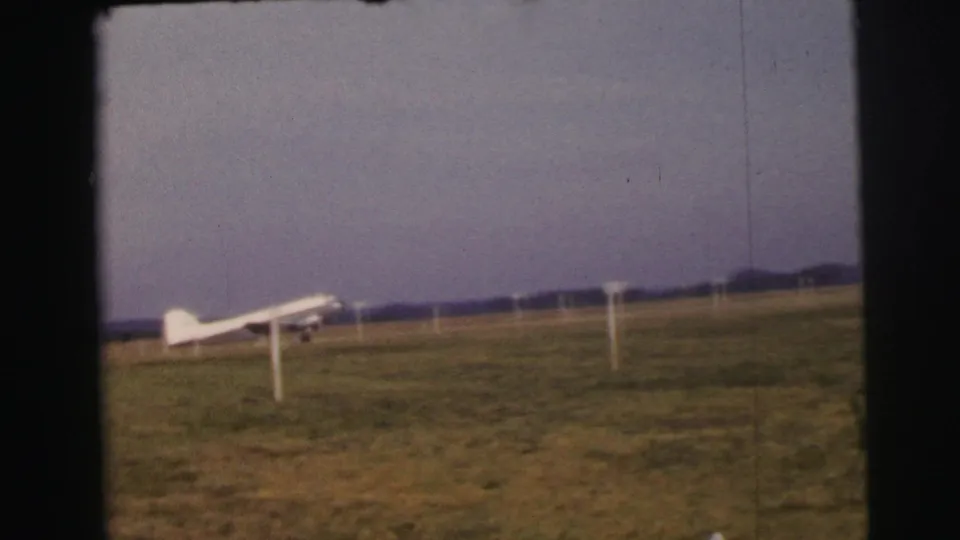 1954 Le Mans home-movie frame showing a small white airplane near a grass runway with white boundary posts.