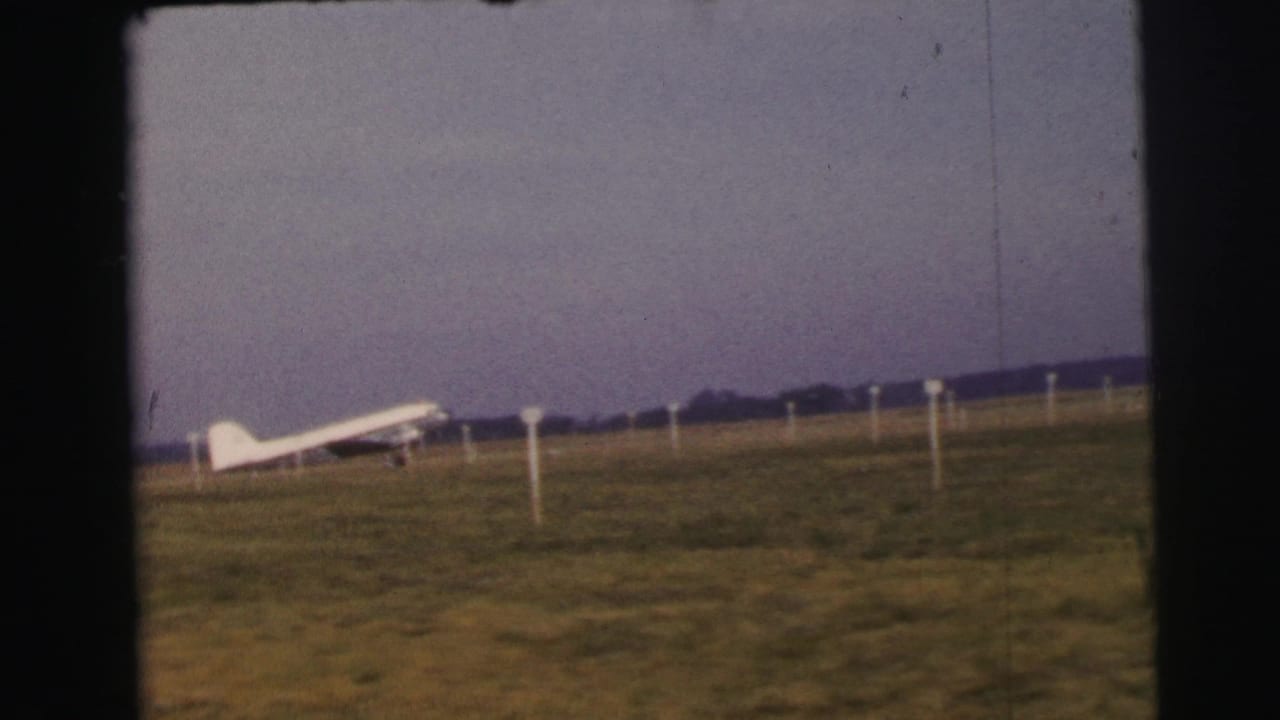 Small white airplane on a Le Mans grass airfield, seen from a distant 8mm home-movie viewpoint.