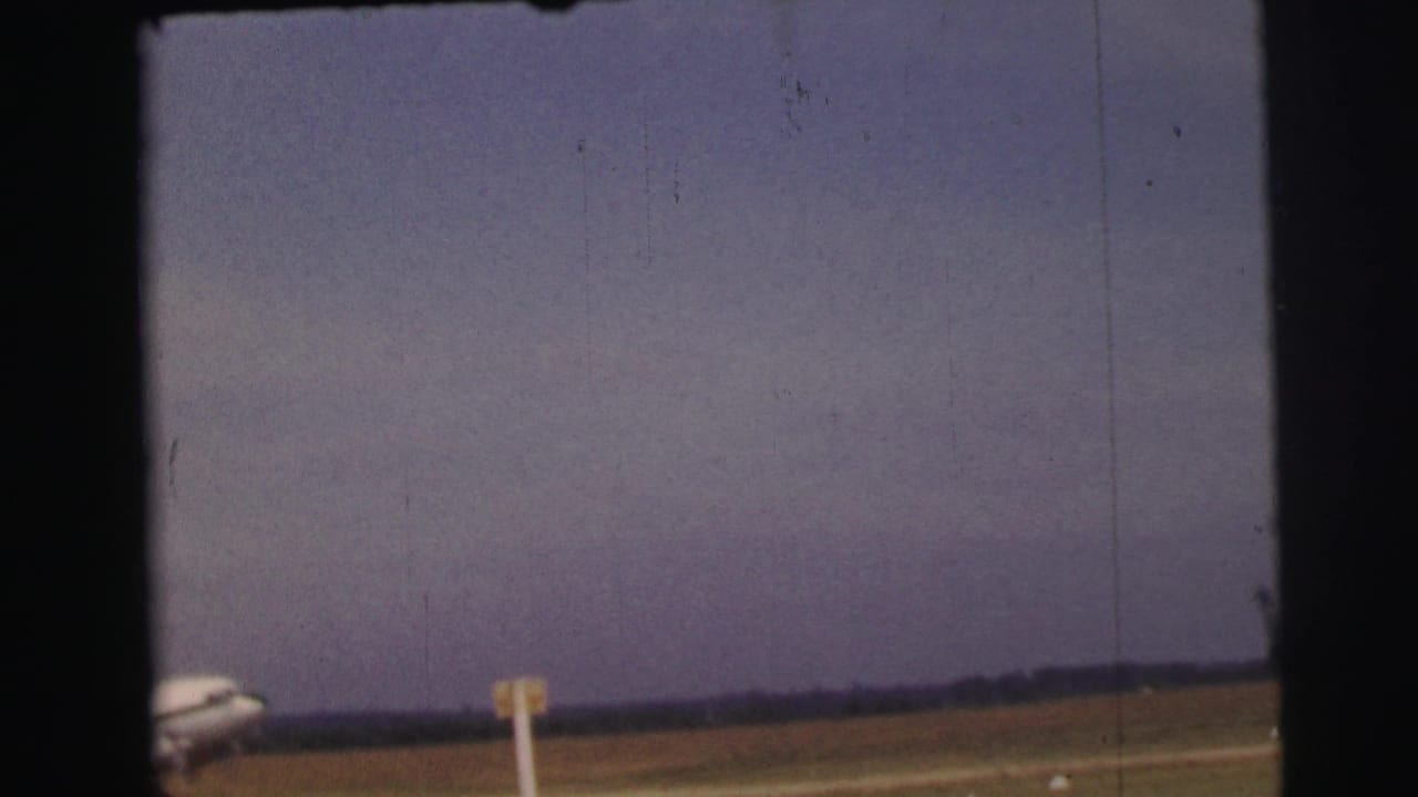 1954 Le Mans home-movie frame showing a distant passenger plane entering view beside a grassy airfield and runway marker.