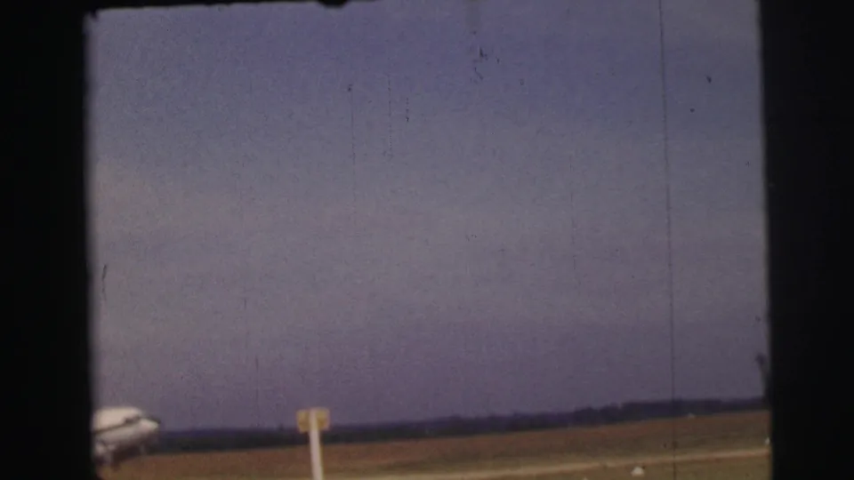 1954 Le Mans home-movie frame showing a distant passenger plane entering view beside a grassy airfield and runway marker.