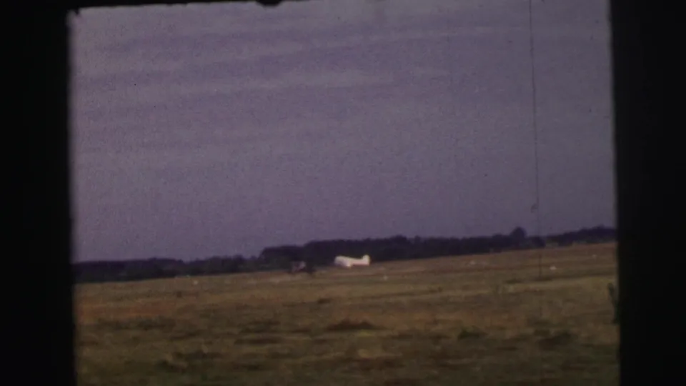 1954 Le Mans home-movie frame with the aircraft now distant across the grass, leaving more of the open airfield visible.