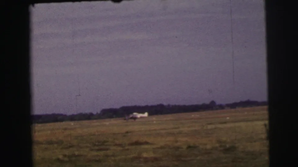 1954 8mm view near Le Mans, France, showing a small aircraft low on a grass airstrip with open field and distant trees.