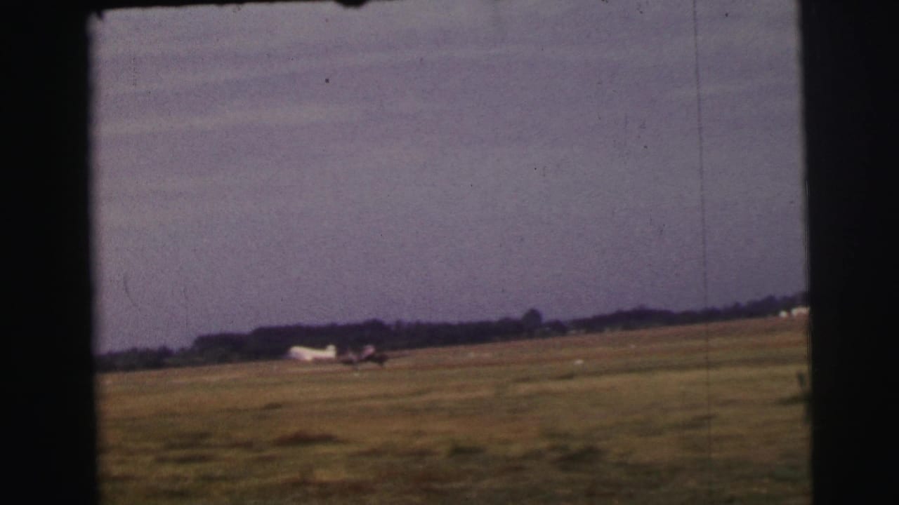 1954 Le Mans home-movie frame of a small aircraft accelerating along a grass runway, seen from a distant amateur viewpoint.