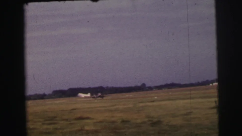 1954 Le Mans home-movie frame of a small aircraft accelerating along a grass runway, seen from a distant amateur viewpoint.