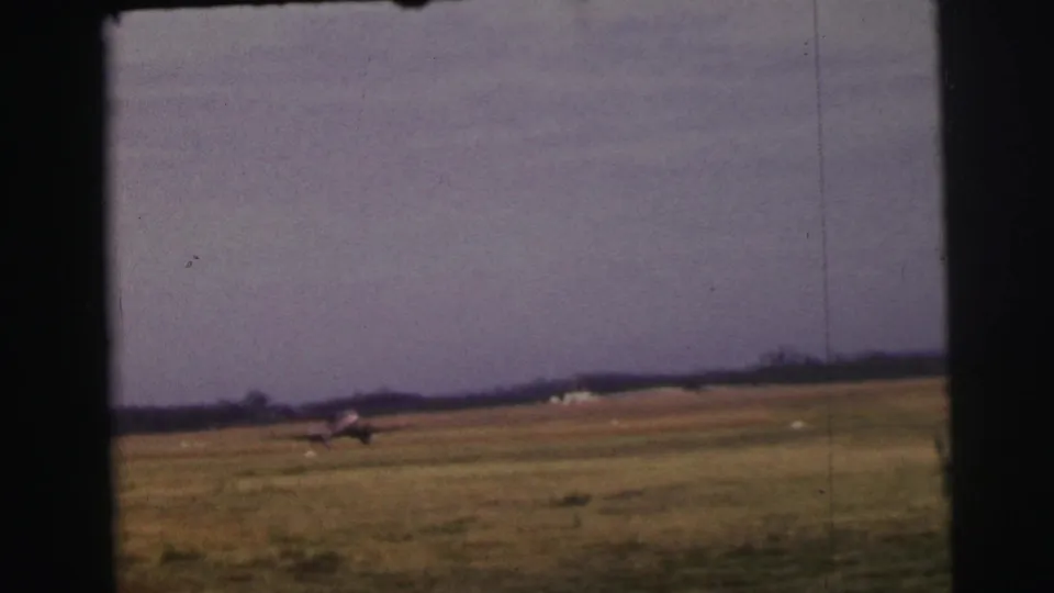 Distant 1954 Le Mans 8mm frame showing a small aircraft on or just above a grass airstrip, with open field and treeline beyond.