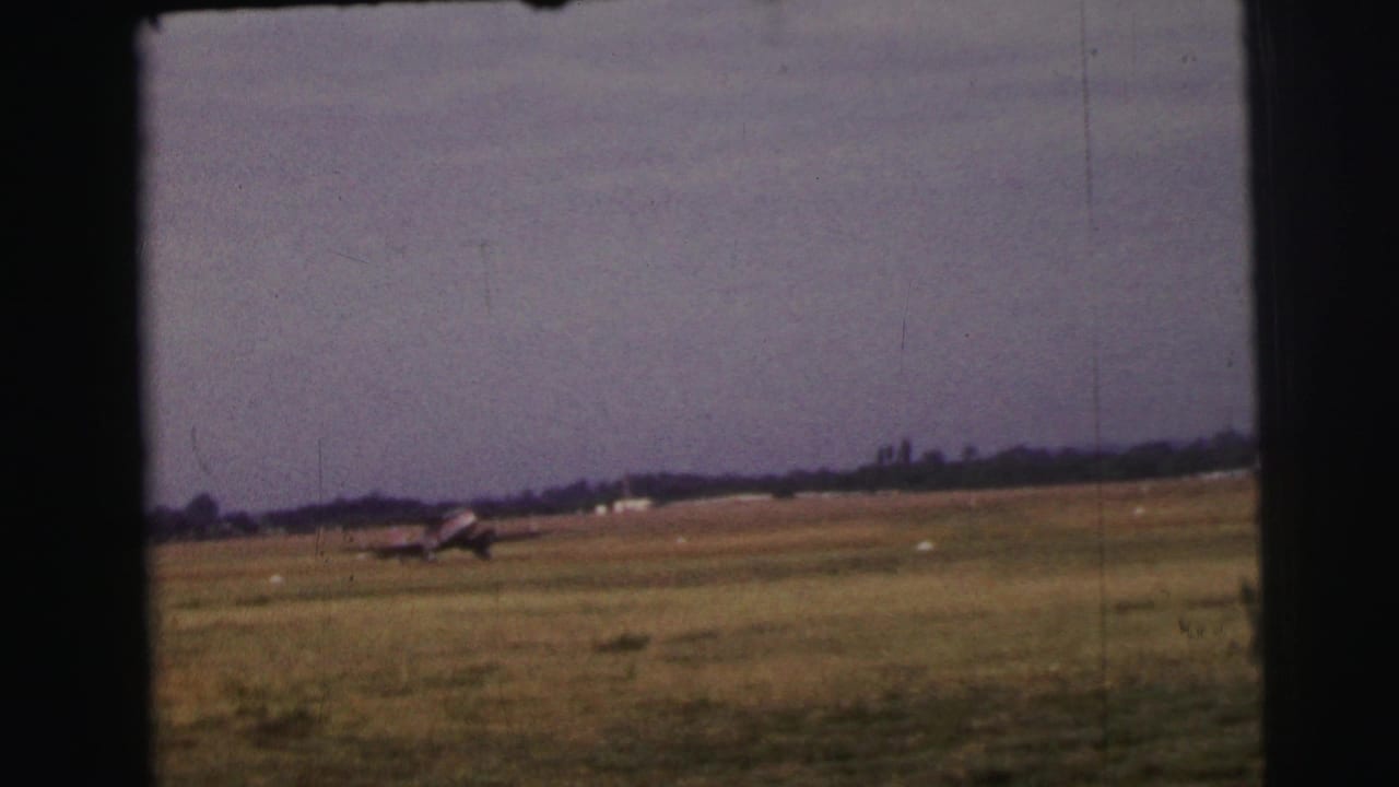 1954 Le Mans home-movie frame showing a small propeller aircraft moving across a grass runway under a pale sky.