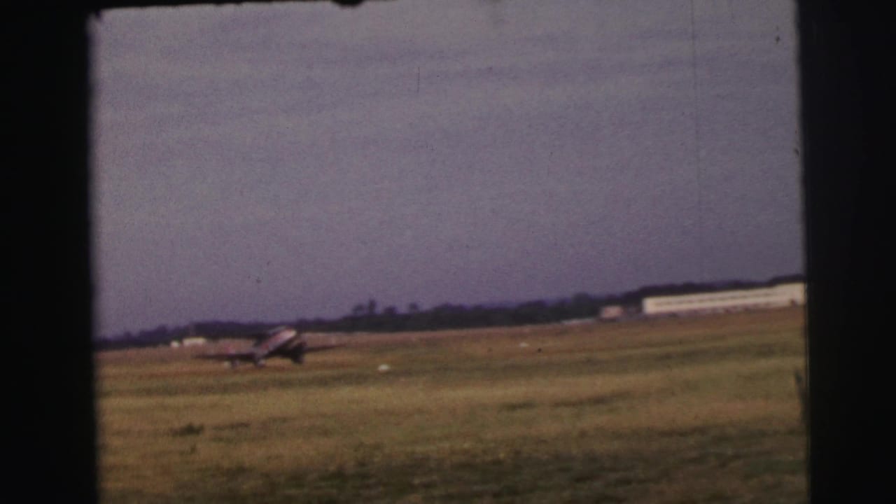 Small aircraft continues across the grass runway at Le Mans, France, with a distant white airfield building visible.
