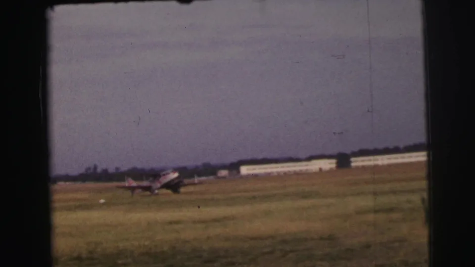 Le Mans 1954 frame of a small propeller plane coasting on grass with a long low airfield structure behind it.