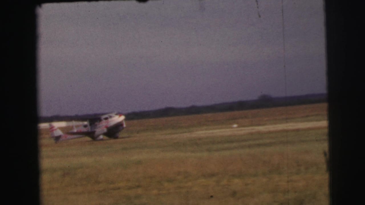 A light-colored propeller aircraft with red markings taxis on the Le Mans grass airfield under an overcast sky.