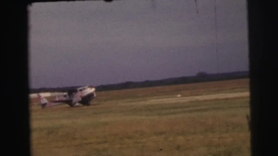 A light-colored propeller aircraft with red markings taxis on the Le Mans grass airfield under an overcast sky.
