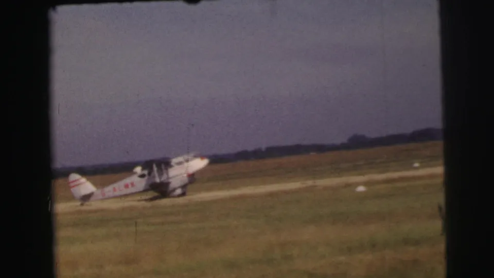 A small propeller airplane appears to slow on the grassy Le Mans airfield, seen in faded 8mm color.