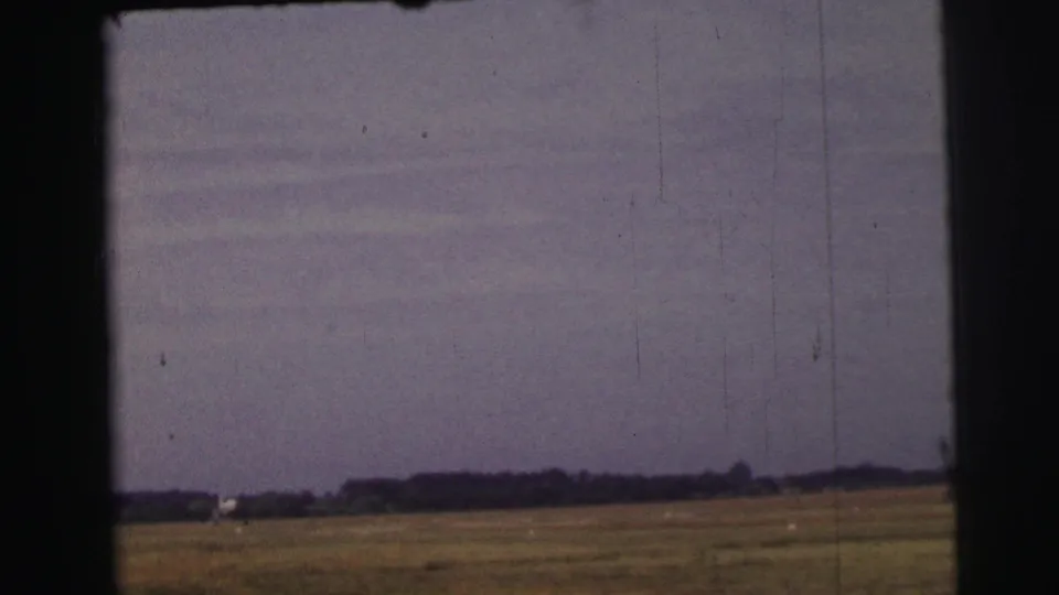 1954 8mm home-movie view near Le Mans showing a flat meadow, distant treeline, pale horizon structure, and dark frame borders.