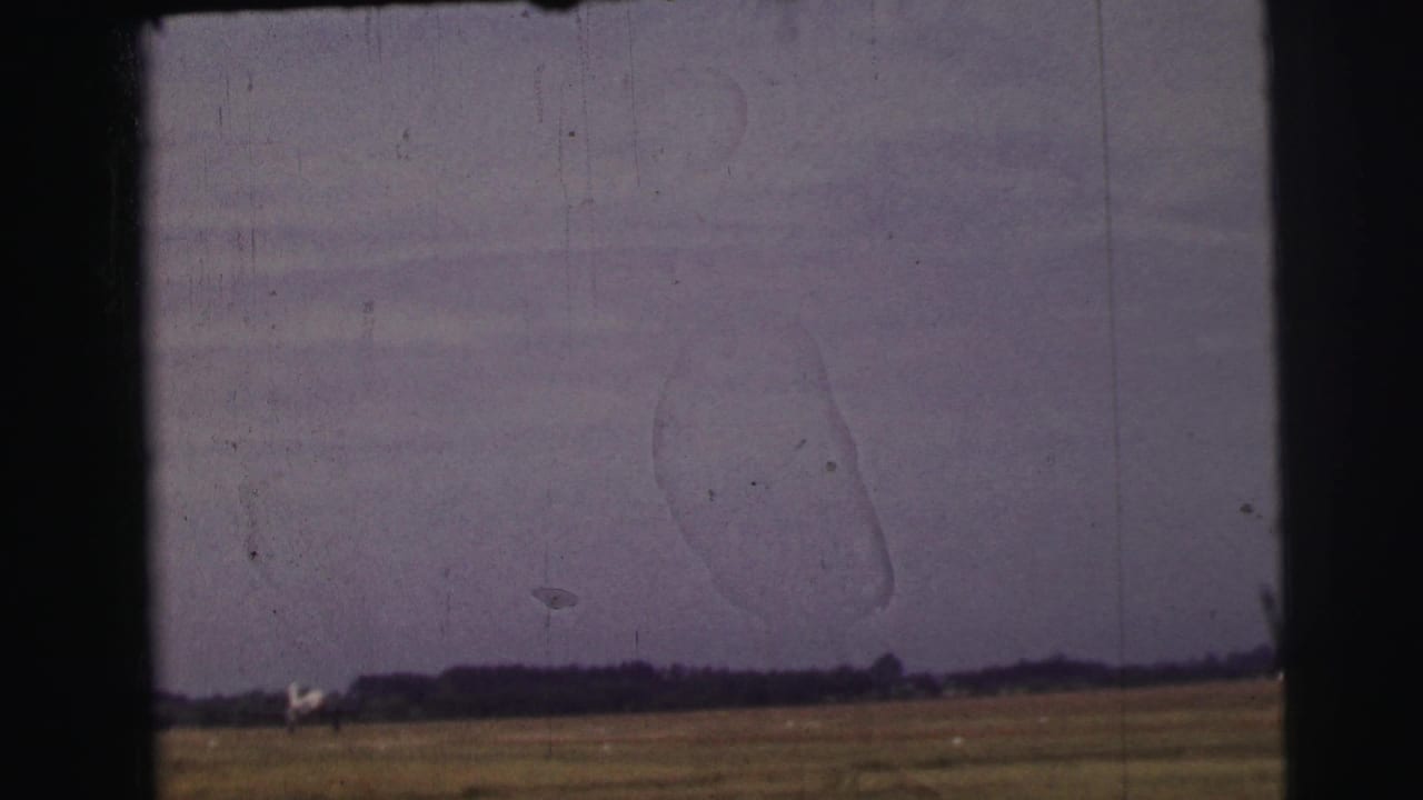 1954 8mm frame near Le Mans with open meadow, low treeline, pale structure on the horizon, and visible archival film wear.