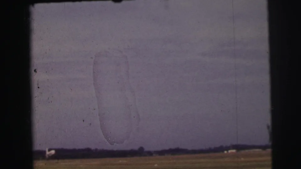 Faded 8mm home-movie view near Le Mans showing a meadow, distant trees, pale horizon shapes, and prominent film surface marks.