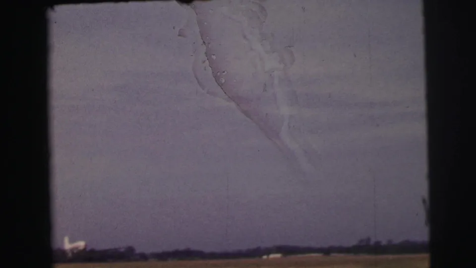 1954 8mm countryside frame near Le Mans with a flat field, distant treeline, small pale structures, dark border, and visible scratches.