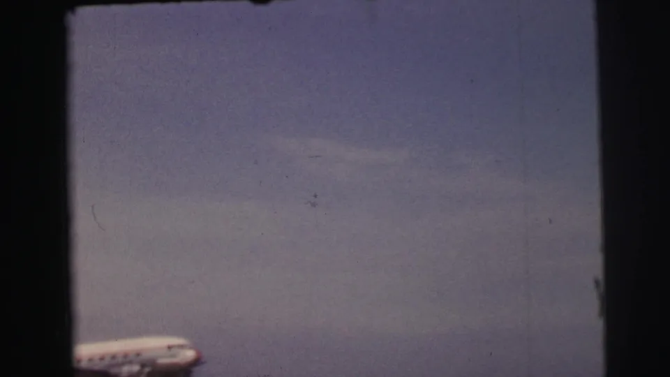 1954 Le Mans home-movie frame showing the blurred nose of a propeller aircraft crossing the lower left against a pale sky.