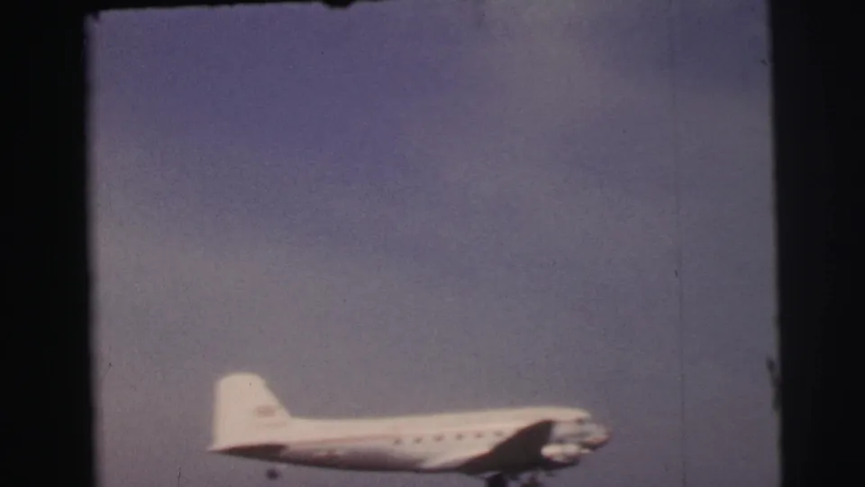 1954 Le Mans 8mm frame showing a light-colored propeller airplane climbing through a hazy sky after takeoff.
