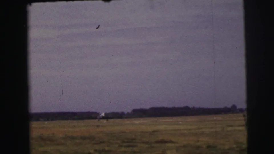 A small aircraft is seen far from the camera across a flat rural field in 1954 Le Mans home-movie footage.