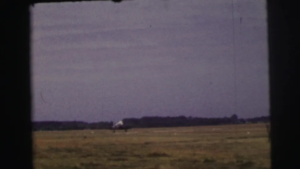 A small propeller airplane appears far across a grassy Le Mans airfield in 1954, framed by dark 8mm film edges.