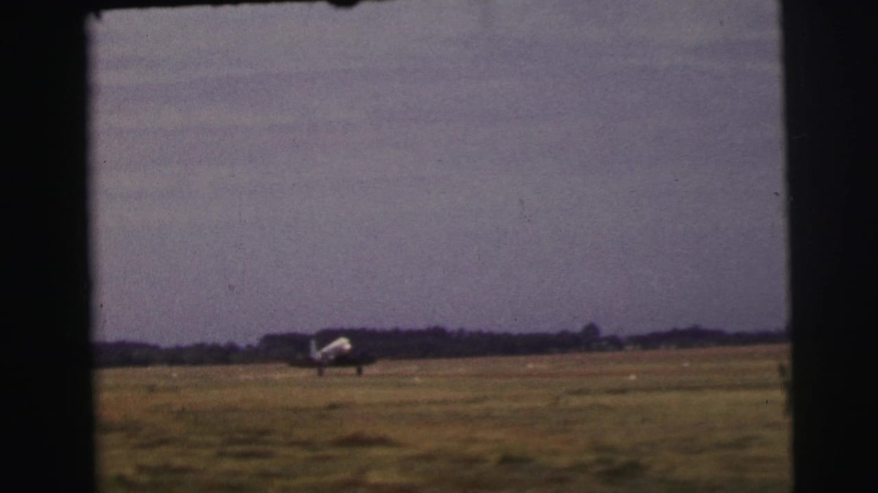 The aircraft taxis across the open grass field at Le Mans, with a low tree line and soft 8mm color visible in the frame.