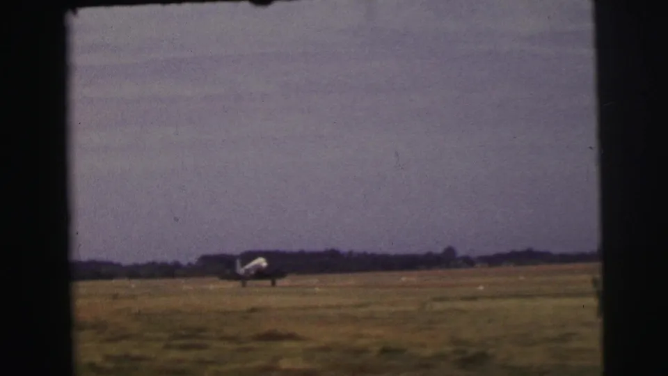 The aircraft taxis across the open grass field at Le Mans, with a low tree line and soft 8mm color visible in the frame.