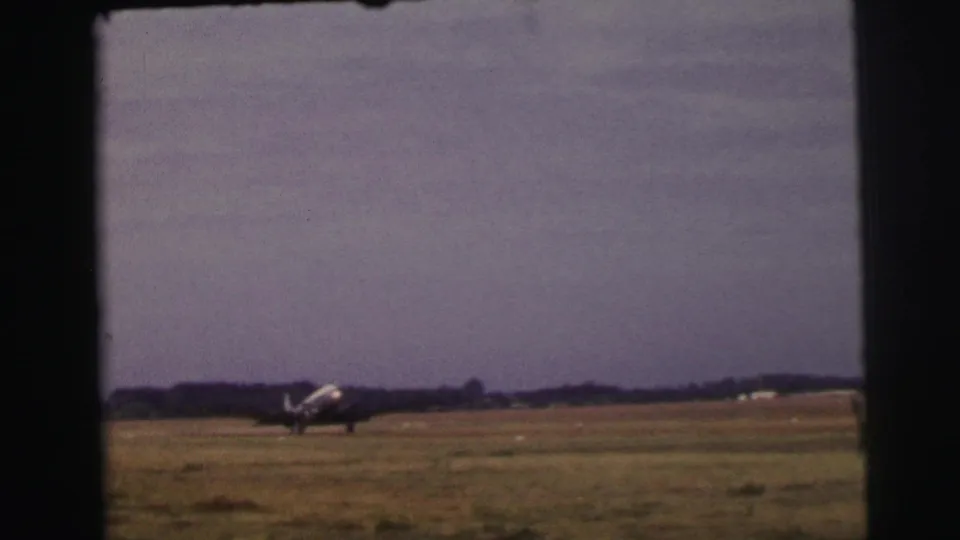 A small mid-century propeller airplane is seen nearer the camera while taxiing over the grass runway in the 1954 Le Mans footage.