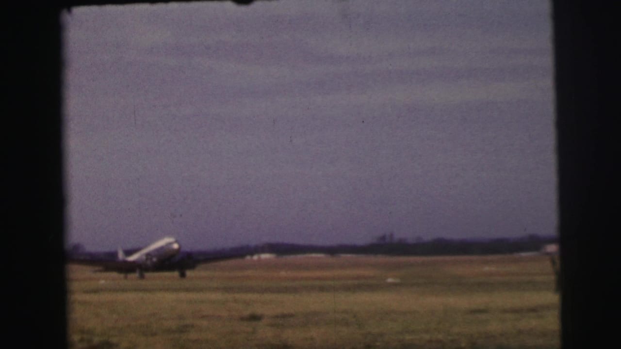 A propeller passenger plane appears at distance on a grassy Le Mans airfield in 1954, framed by dark 8mm film edges.