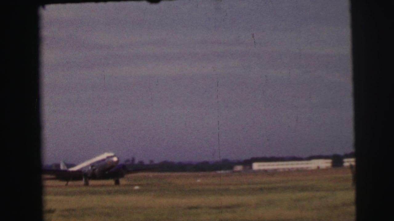 The silver propeller airliner moves across the grass airfield at Le Mans, with low buildings visible in the distance.