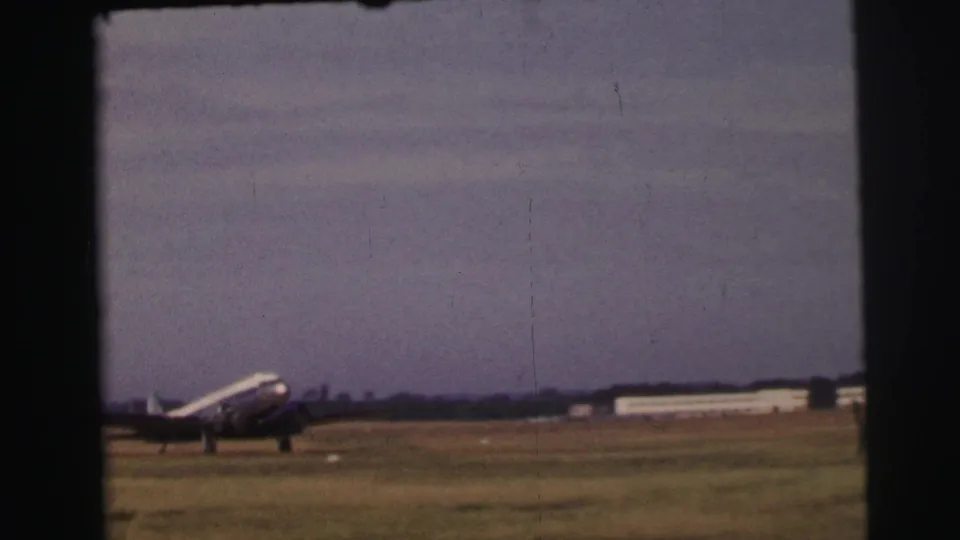 The silver propeller airliner moves across the grass airfield at Le Mans, with low buildings visible in the distance.
