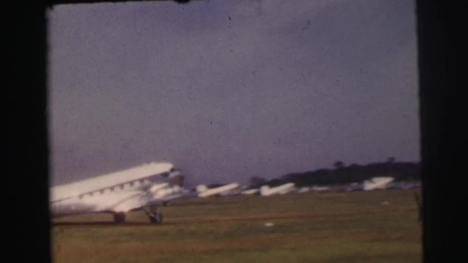 The propeller aircraft exits the frame while small parked planes appear beyond the grassy runway at Le Mans.