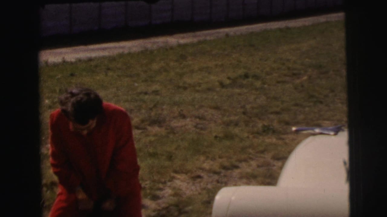 At Le Mans, France in 1954, a woman in a red dress bends near a pale parked car on grass, with the frame partly darkened by film borders.