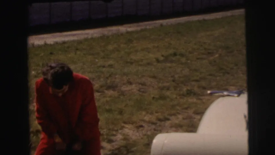 At Le Mans, France in 1954, a woman in a red dress bends near a pale parked car on grass, with the frame partly darkened by film borders.