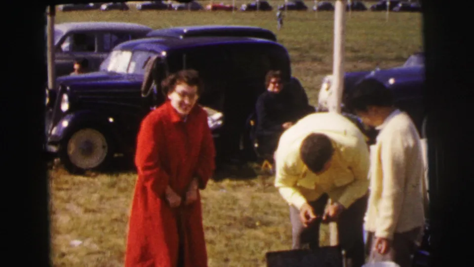 At Le Mans, France in 1954, a woman in red faces the camera near a man bending over a case, with dark vintage cars parked behind them.