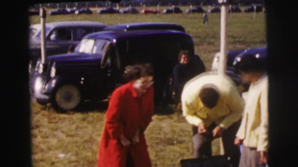 At Le Mans, France in 1954, the woman in red moves past spectators near a white post, a dark sedan, and parked cars on the grassy field.