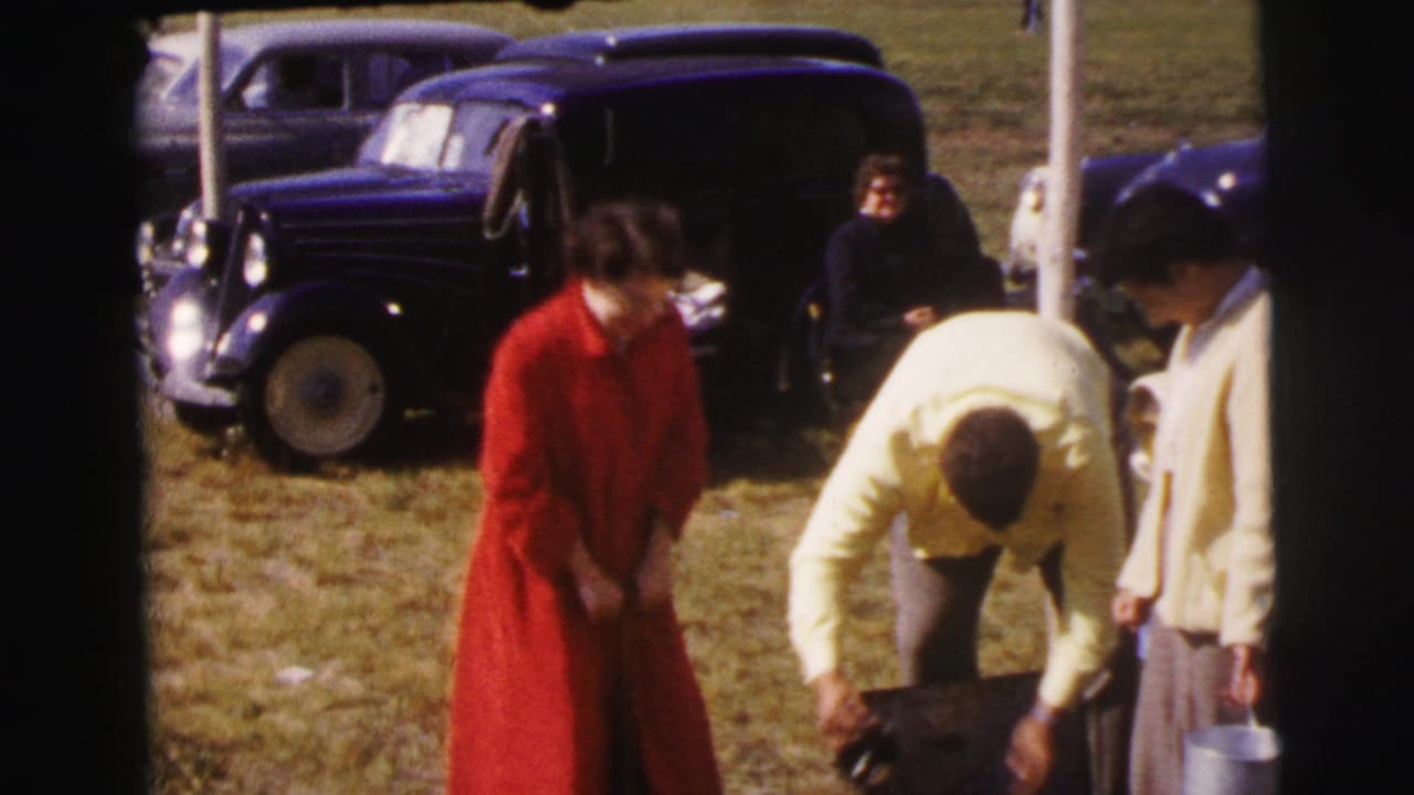 Adults stand beside dark vintage cars on a grassy field, with a woman in a red coat and a man bending near a bag.