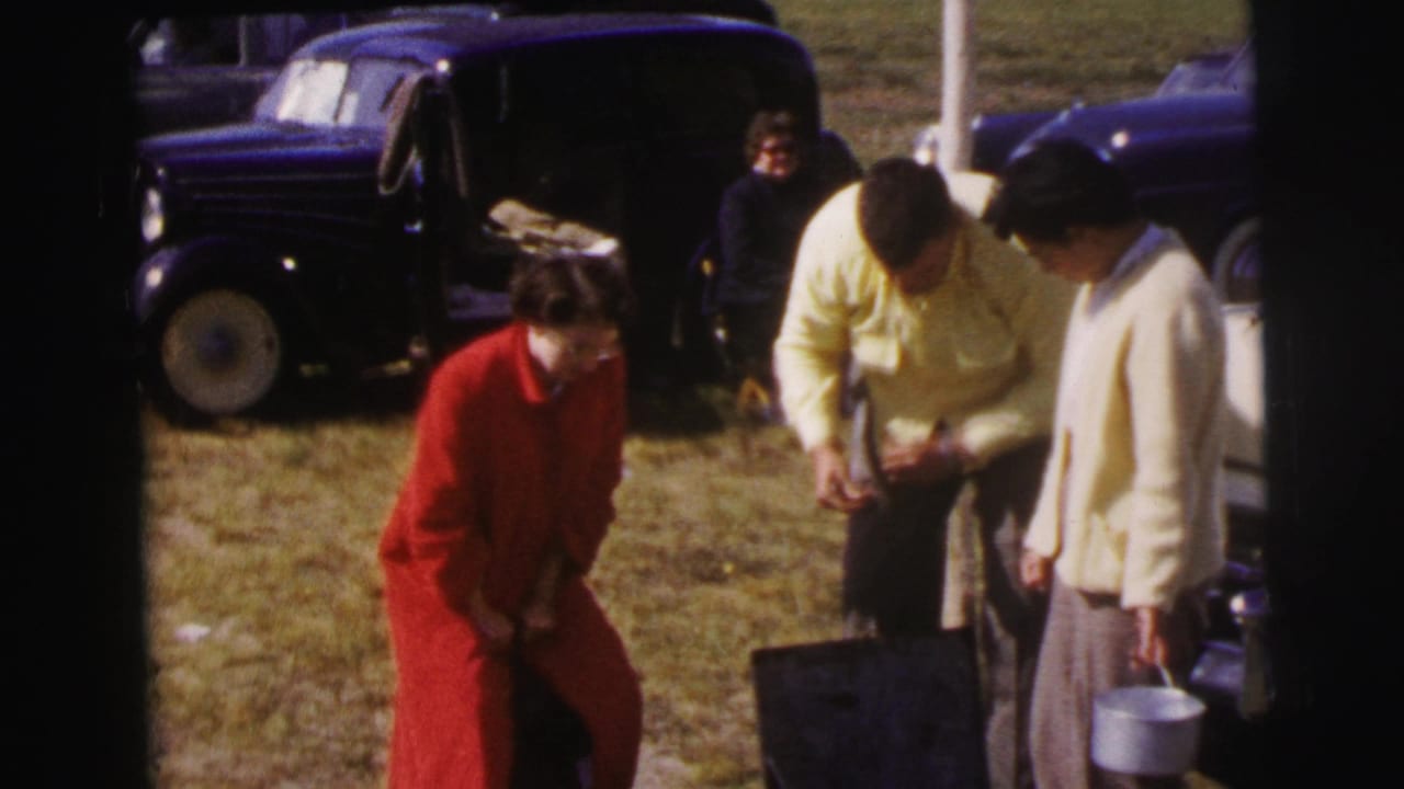 At the 1954 Le Mans setting, two women stand near parked cars while a man bends over a case and another person sits in the background.