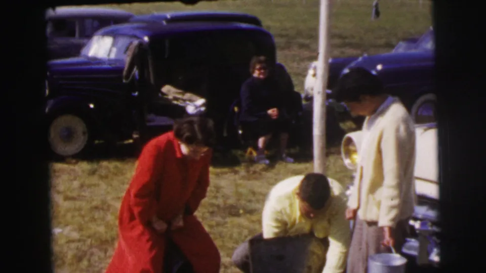 In 1954 Le Mans home-movie footage, a woman in red leans forward while a man crouches near a case and a woman holds a metal pail.