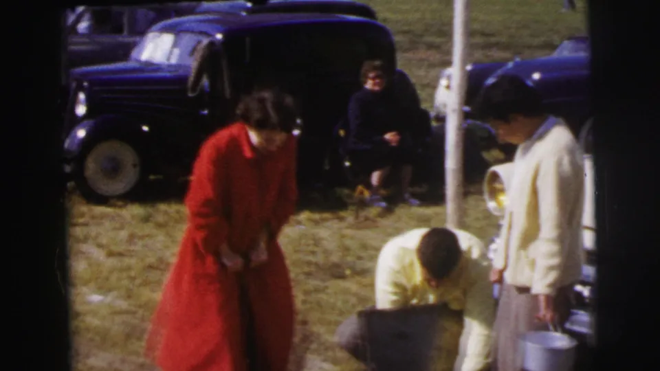 At a 1954 Le Mans roadside stop, a woman in red stands foreground as a man crouches by a case and another woman waits beside a car.