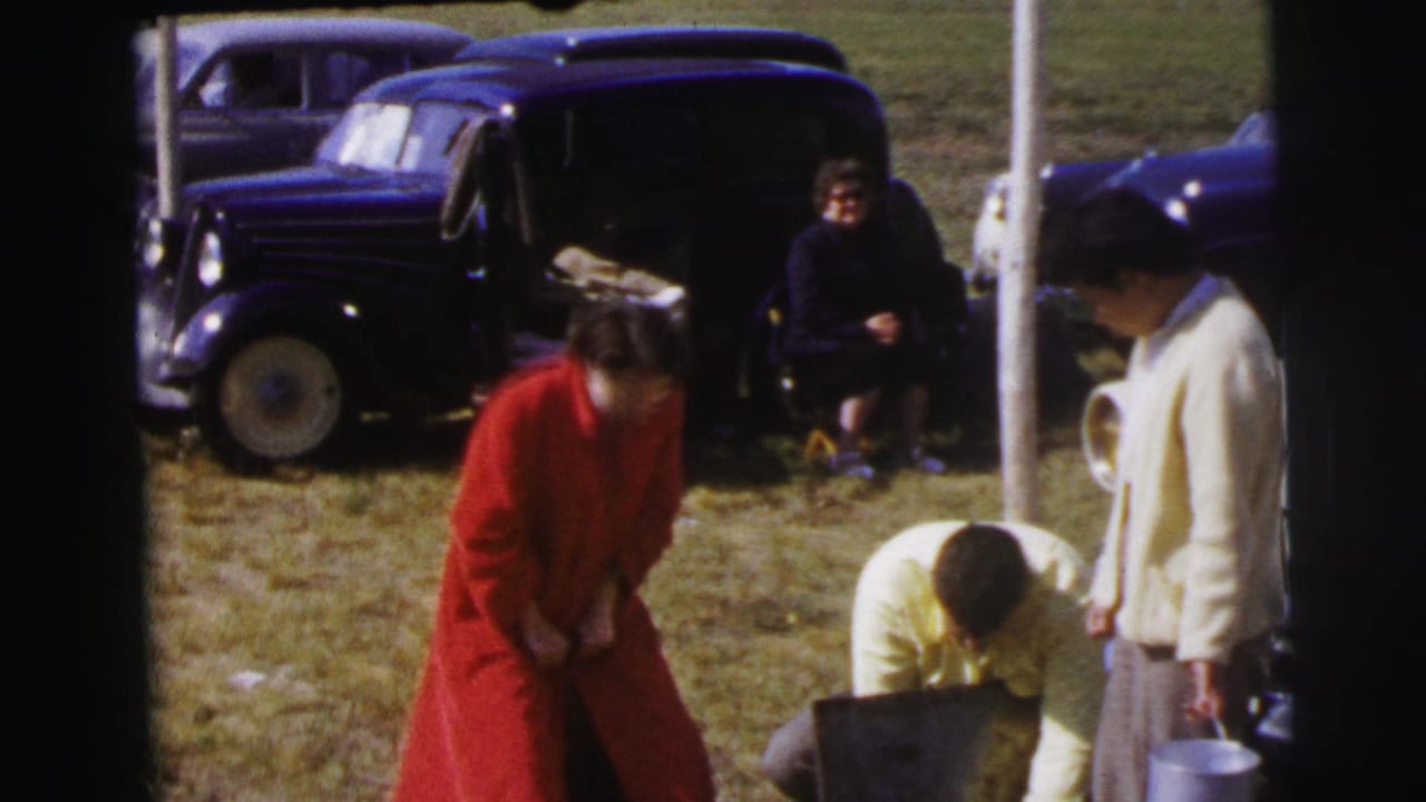 At Le Mans, France in 1954, a woman in a red coat stands near a crouching man while mid-century cars are parked on grass.