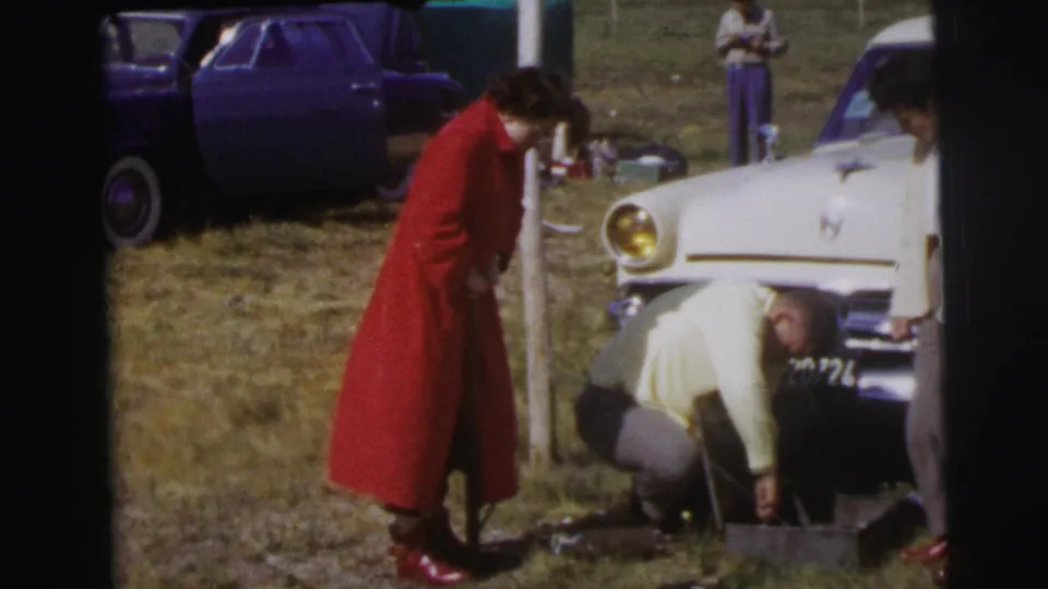 In the 1954 Le Mans home-movie footage, a man crouches at the front of a white car as the woman in red observes.
