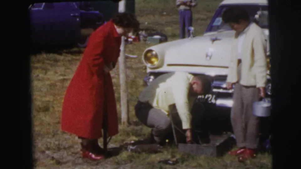 At Le Mans in 1954, the man remains crouched by the white car's front bumper while two women stand beside the roadside repair scene.